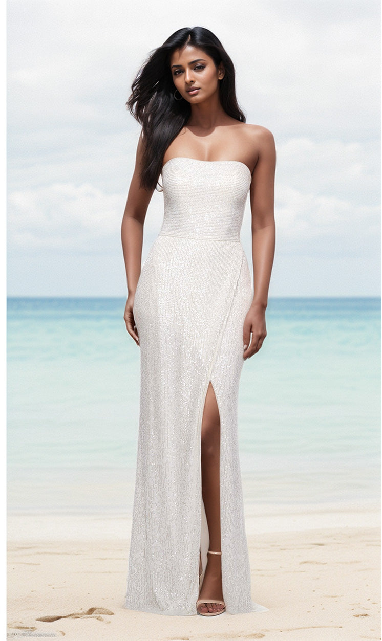 A woman stands on a sandy beach in the white La Femme Strapless Long Sequin Prom Dress 29681, featuring a thigh-high slit and shimmering sequins that catch the light with the ocean in the background.